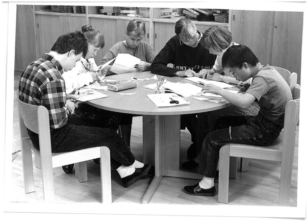 5 Kinder sitzen an einem Tisch und lernen. Eine Frau sitzt dabei und hilft ihnen.