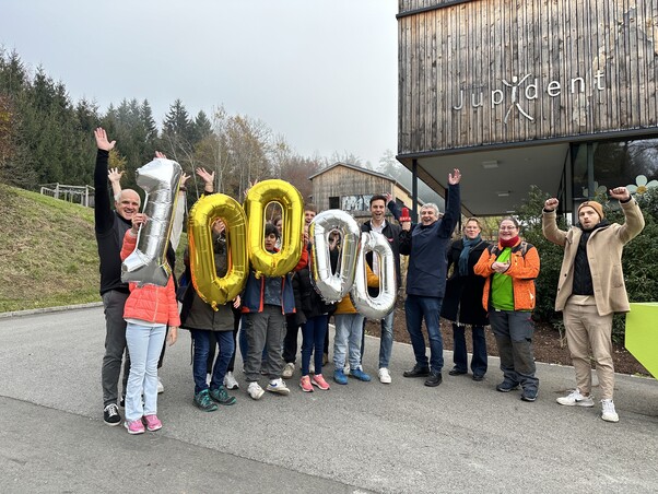 Gruppenfoto mit vielen Personen. Sie halten Luftballons mit der Zahl 10.000 in die Höhe. Sie freuen sich