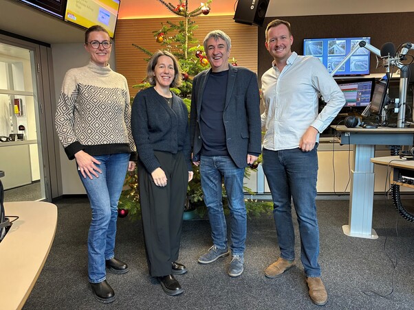 2 Frauen und 2 Männer stehen in einem Tonstudio. Im Hintergrund ein Weihnachtsbaum. Sie lächeln alle.
